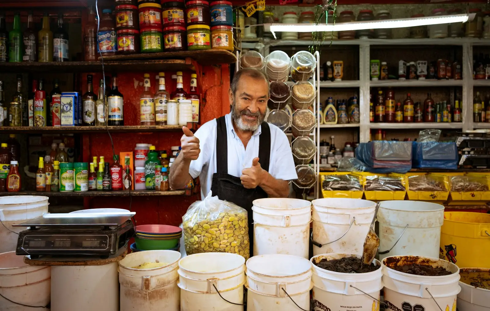 Dueño de un puesto de abarrotes, sonriendo con el pulgar arriba detrás del mostrador.